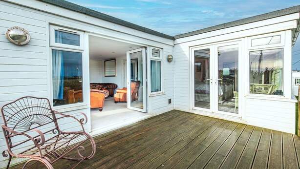 A covered outdoor area with a wooden deck, a metal chair, and a view into a room with a sofa and curtains.
