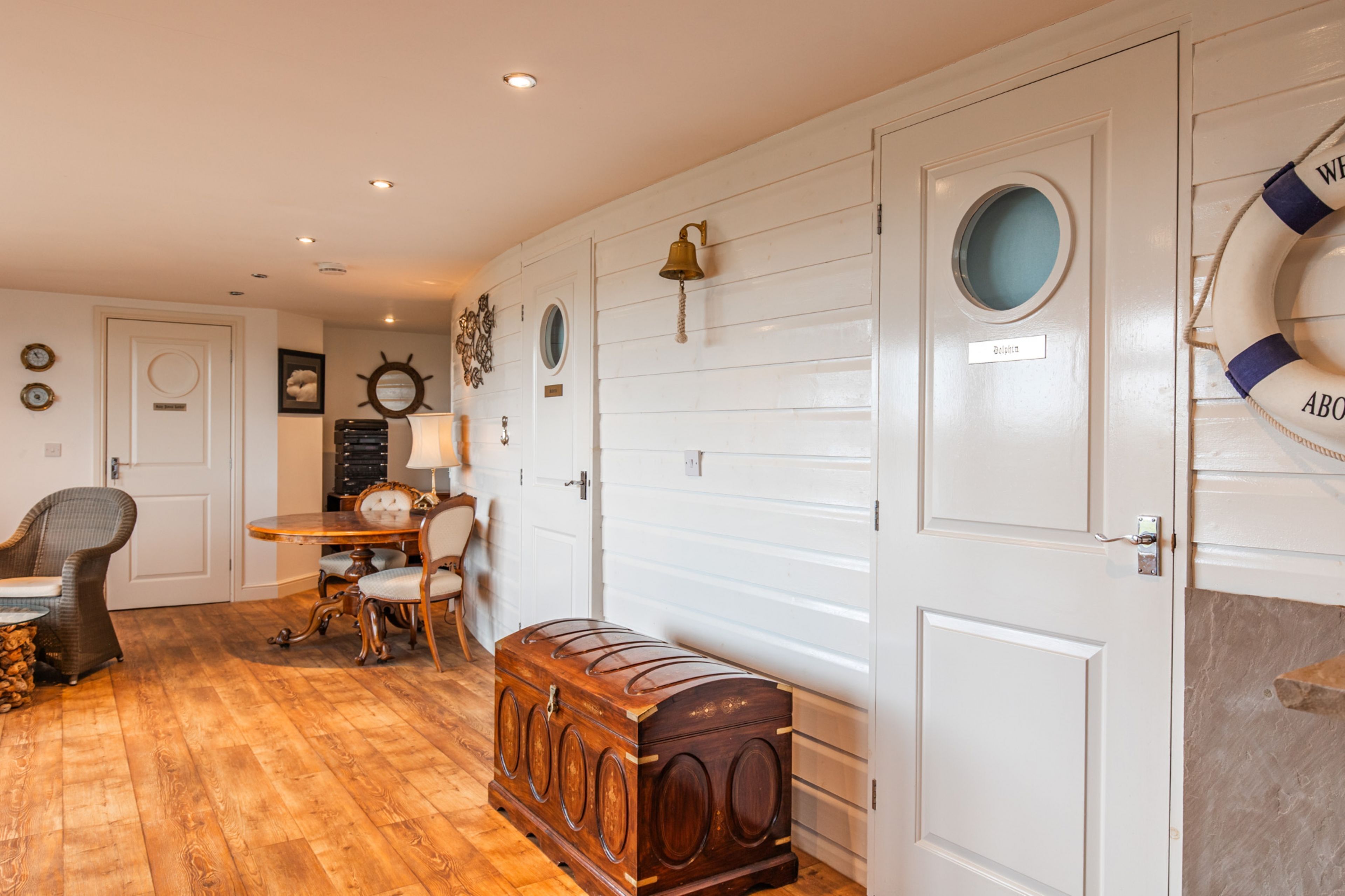 A room with a wooden floor, a dining table with chairs, a small wooden chest, and a life preserver on the wall.