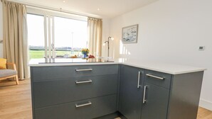A modern kitchen with grey cabinets, a white countertop, and a view of a green landscape through large windows. - Sea Fern House - 36 (Poole)