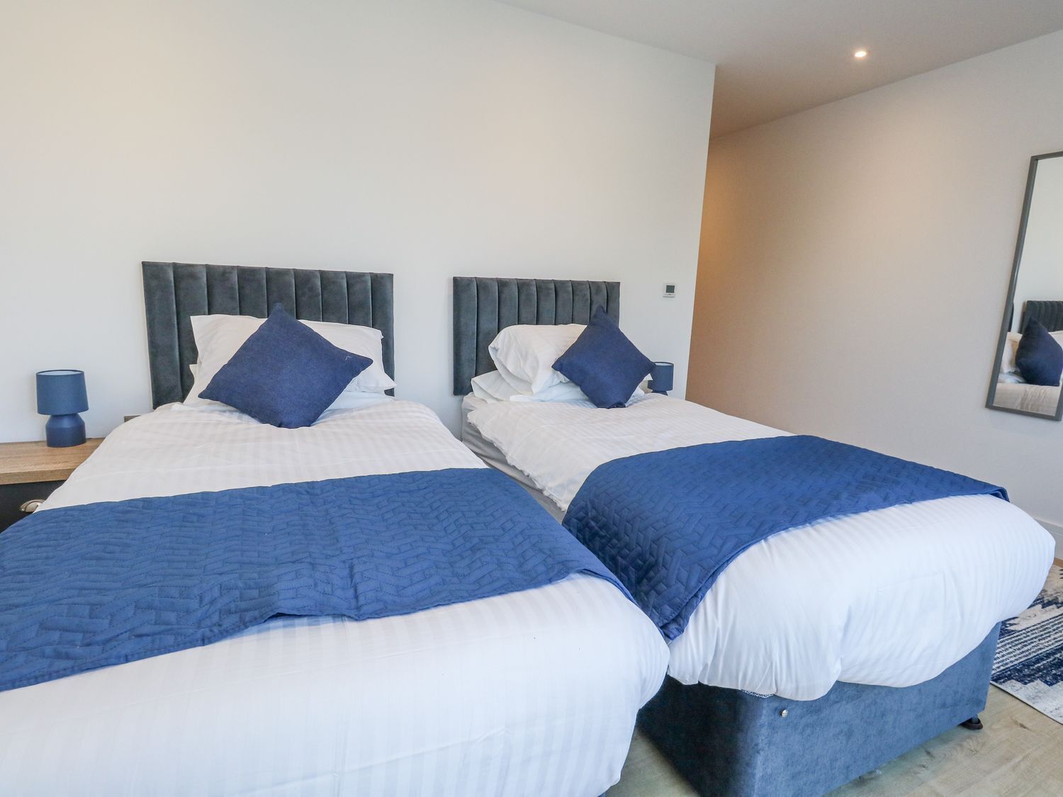 Een hotelkamer met twee bedden, elk met een grijze hoofdbord en wit beddengoed. Er staat een klein tafeltje met een lamp naast elk bed.