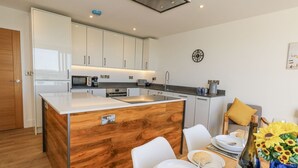 A modern kitchen with white cabinets, a wooden island, and a dining area with a table set for two. - Sea Fern House - 36 (Poole)