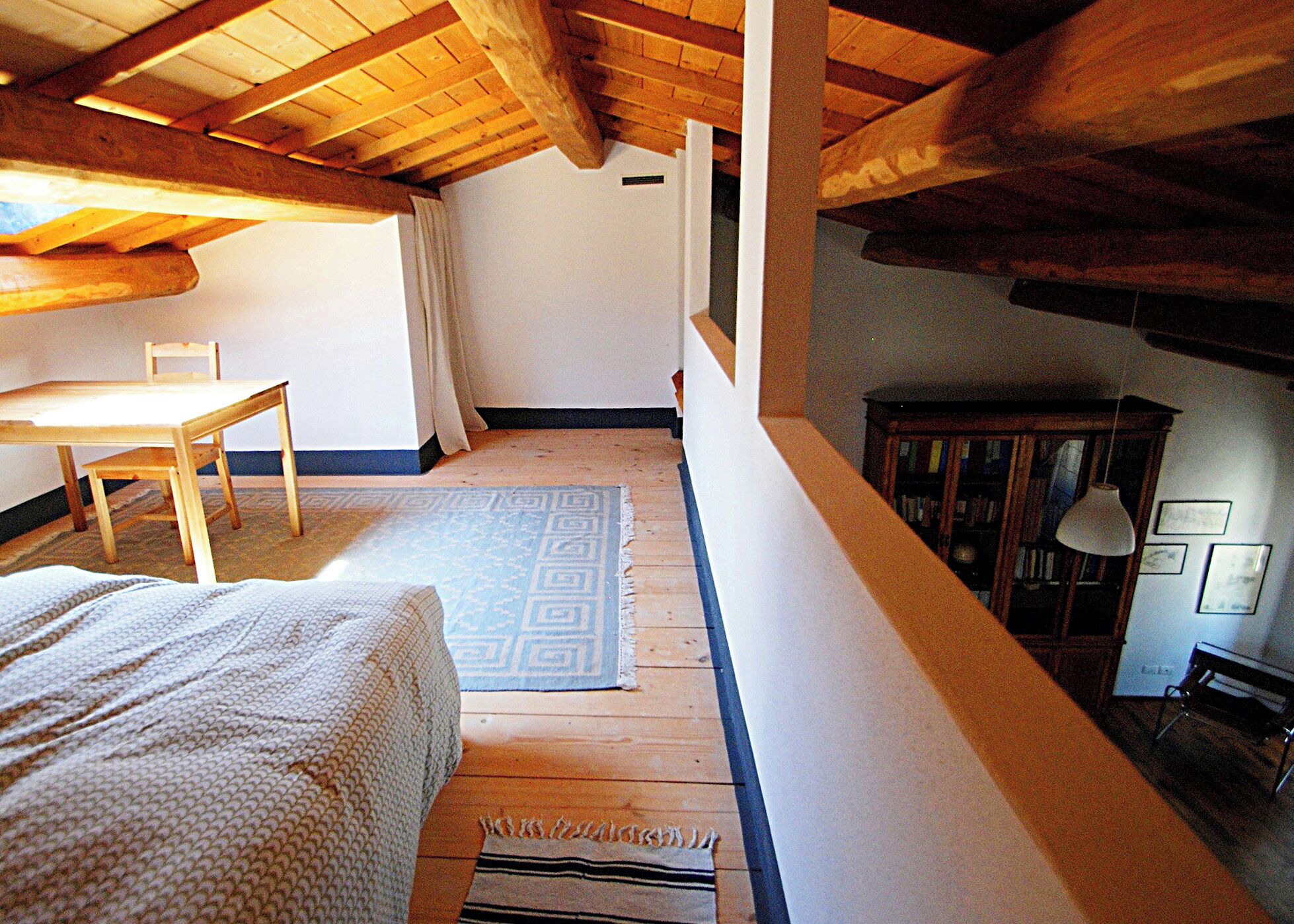 Quarto com teto de madeira, uma cama, uma mesa e uma cadeira. Há um tapete no chão e um armário com livros.