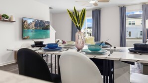 Uma sala de jantar com mesa posta para dois, um ventilador de teto, uma TV na parede e vista para uma casa pela janela.