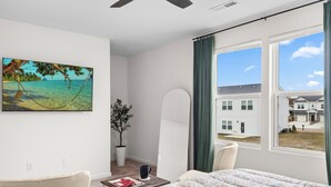 Quarto com cama, ventilador de teto, janela com vista para casas, uma planta e um quadro de uma praia.