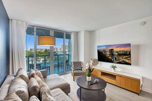 A modern living room with a sofa, a round coffee table, a TV, and a view of a cityscape through large windows. - Stunning 1-Bedroom Apartment Steps from the Ocean (Hallandale Beach)