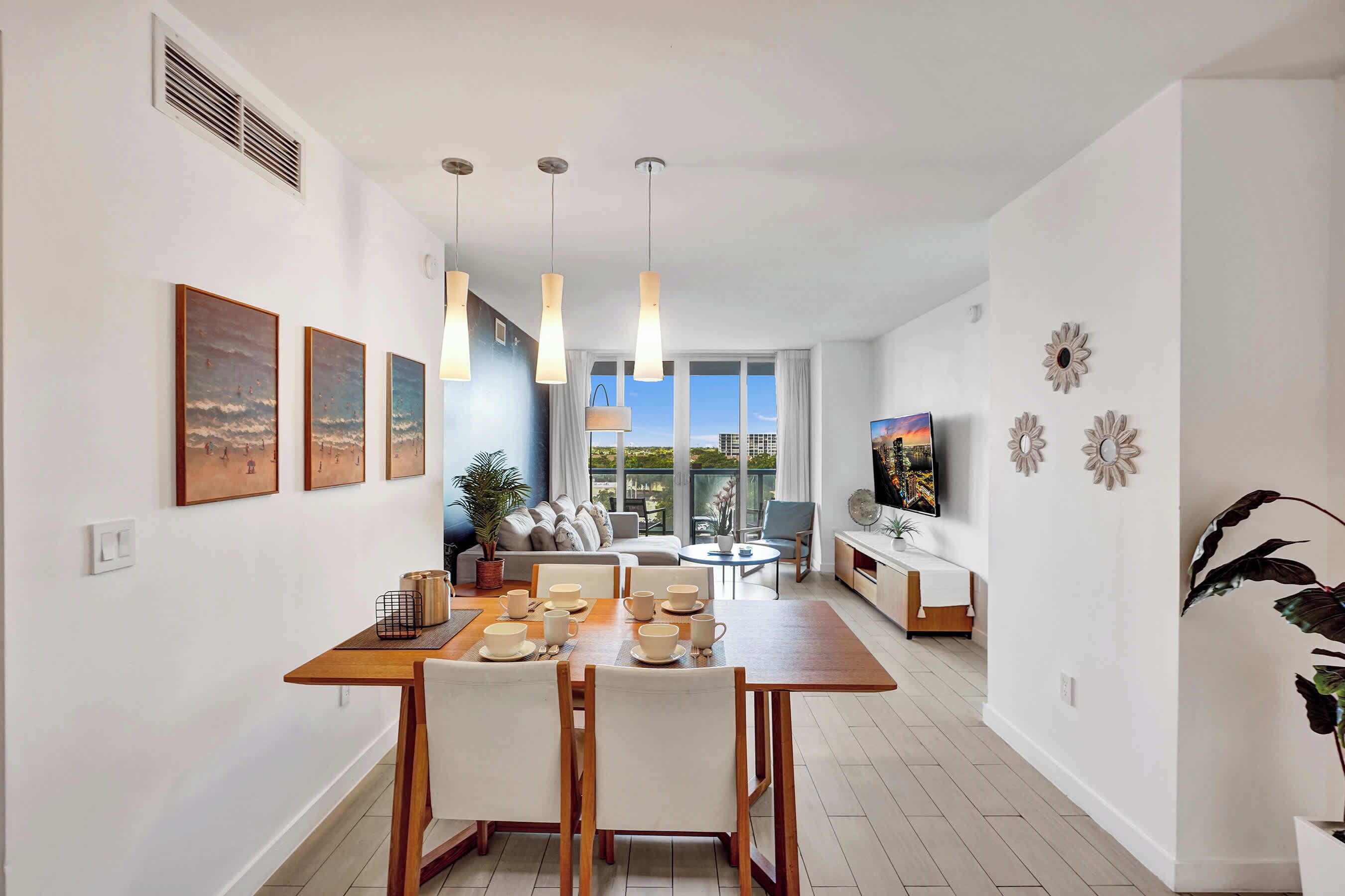 A modern living room with a dining area, large windows, and a flat-screen TV mounted on the wall.
