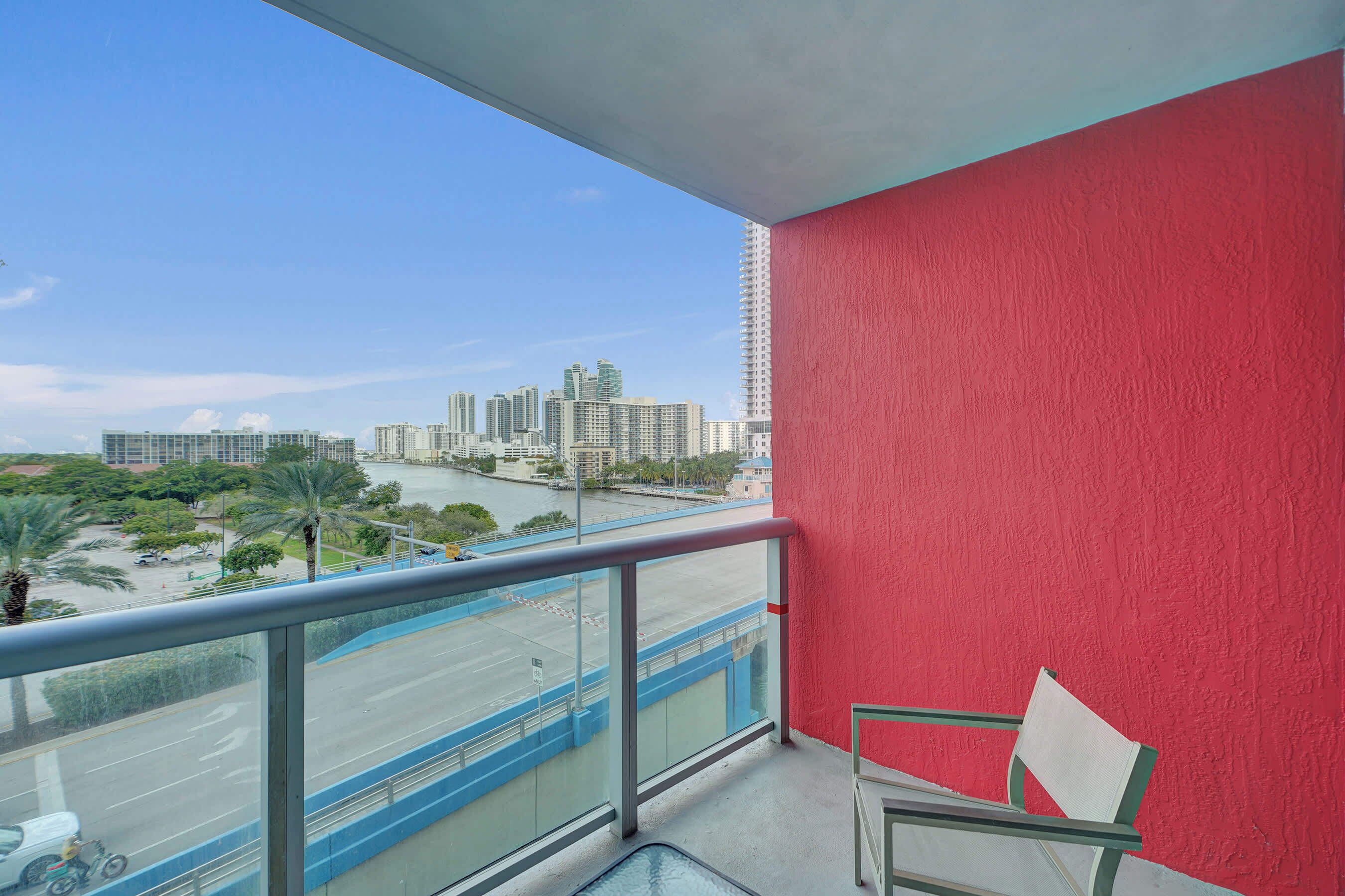 Un balcón con pared roja, barandilla de vidrio y vista a un paisaje urbano con rascacielos y palmeras.