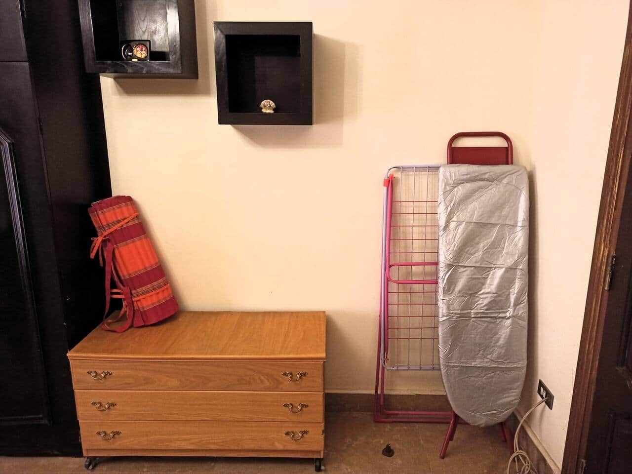 Une petite commode en bois, sur laquelle est posé un objet à carreaux rouges, une étagère noire avec une horloge, et un étendoir métallique recouvert d’une housse blanche.