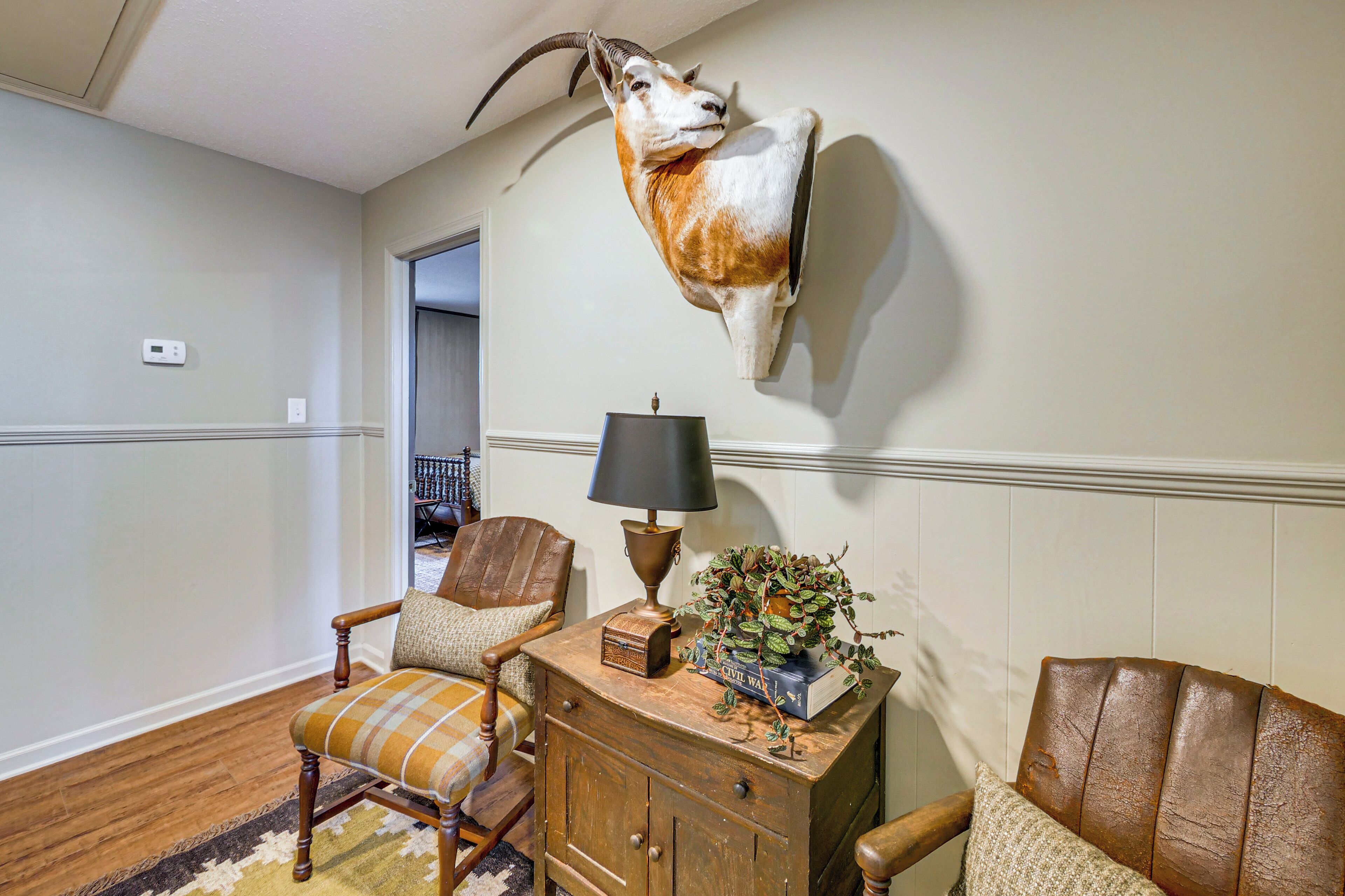 A room with a mounted animal head, a wooden cabinet, a lamp, and two chairs.