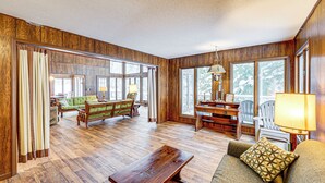 A spacious living room with wooden floors, a green sofa, a wooden coffee table, and a large window providing natural light. - Near Manistee National Forest! Retro Cabin Retreat (Baldwin)