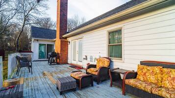 Une terrasse en bois avec des meubles d’extérieur, une cheminée en briques et une maison blanche avec une porte verte.