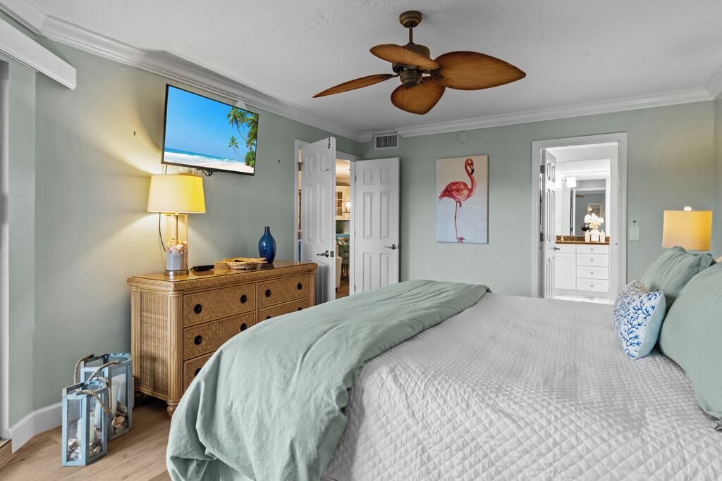 Un dormitorio con una cama, un ventilador de techo, un televisor, un aparador y una puerta que da a otra habitación.