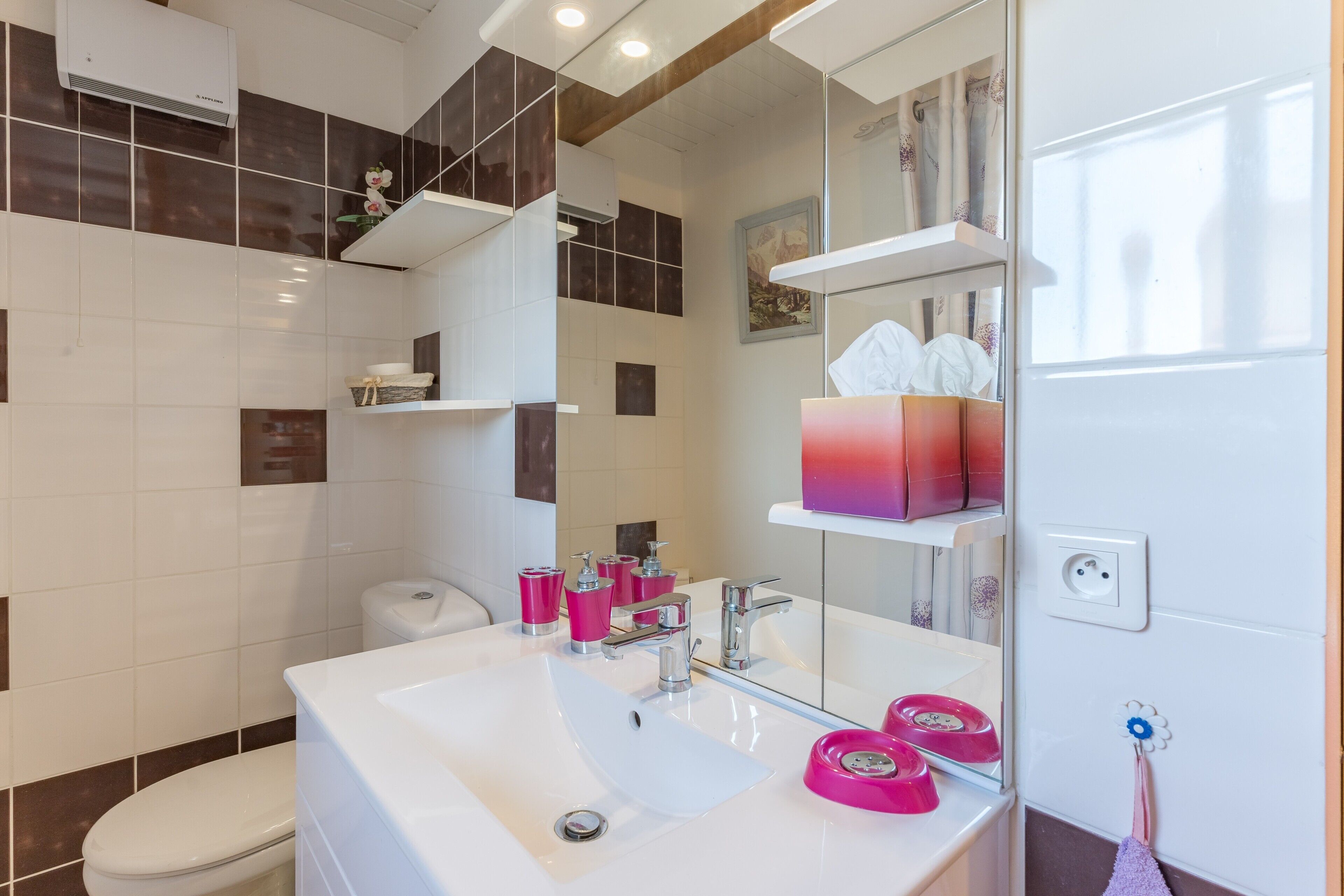 A modern bathroom with a white sink, mirror, and a toilet. There are shelves with toiletries and a towel rack.