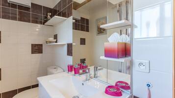 A modern bathroom with a white sink, mirror, and a toilet. There are shelves with toiletries and a towel rack.