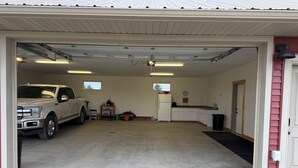 Een garage met een witte pick-up truck erin, een klein keukentje met een koelkast en een raam, en een deur naar een overdekt buitengebied.