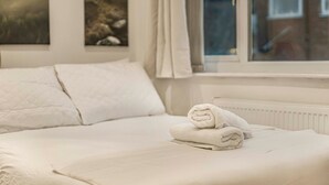 Ein ordentlich bezogenes Bett mit weißen Leinen und einem aufgerollten Handtuch in einem Zimmer mit Fenster und Heizkörper.