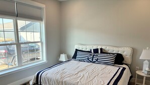 A bedroom with a large bed, a window with blinds, a bedside table with a lamp, and a view of parked cars outside. - Perfect 3-bedroom house with AC in soothing Waterview @ Baypoint (Lakeside Marblehead)