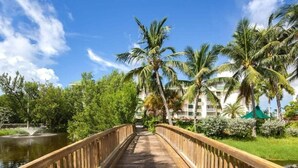 A wooden bridge over a body of water, surrounded by lush greenery and palm trees, leading to a building with a green roof. - The Grand Turk | Spectacular Sunset View, Private (Key West)