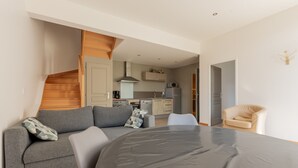 A modern living room with a grey sofa, a dining table, and a kitchen in the background. - Holiday Home 'Gite Clémentine' with Private Terrace, Shared Garden and Wi-Fi (Baromesnil)