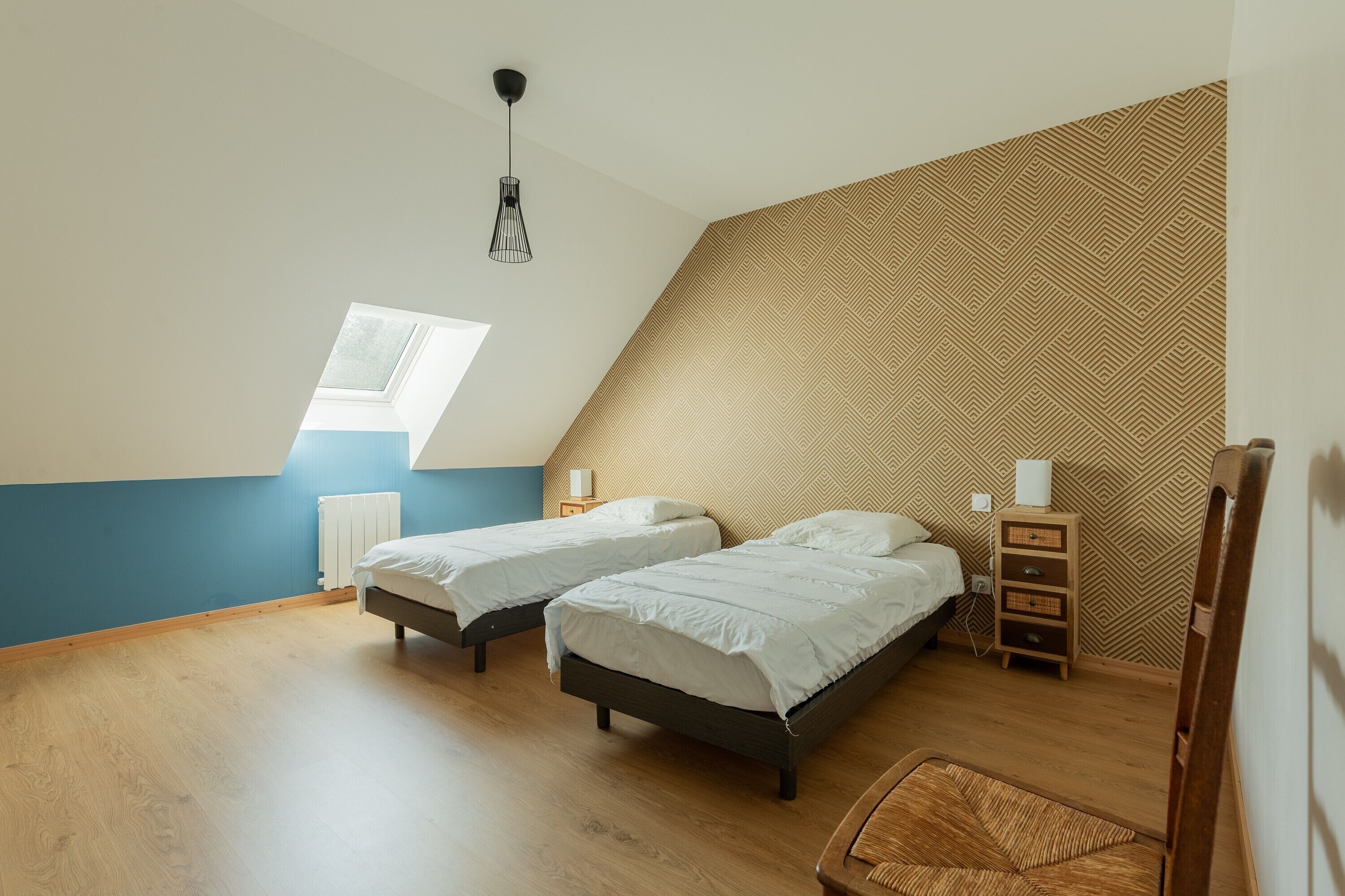 Une chambre avec deux lits, une fenêtre, un radiateur, une commode en bois et un luminaire suspendu.