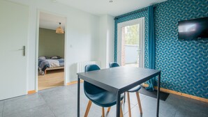 Ein modernes Zimmer mit Esstisch, Stühlen, einem an der Wand montierten Fernseher und einem sichtbaren Bett im angrenzenden Raum.