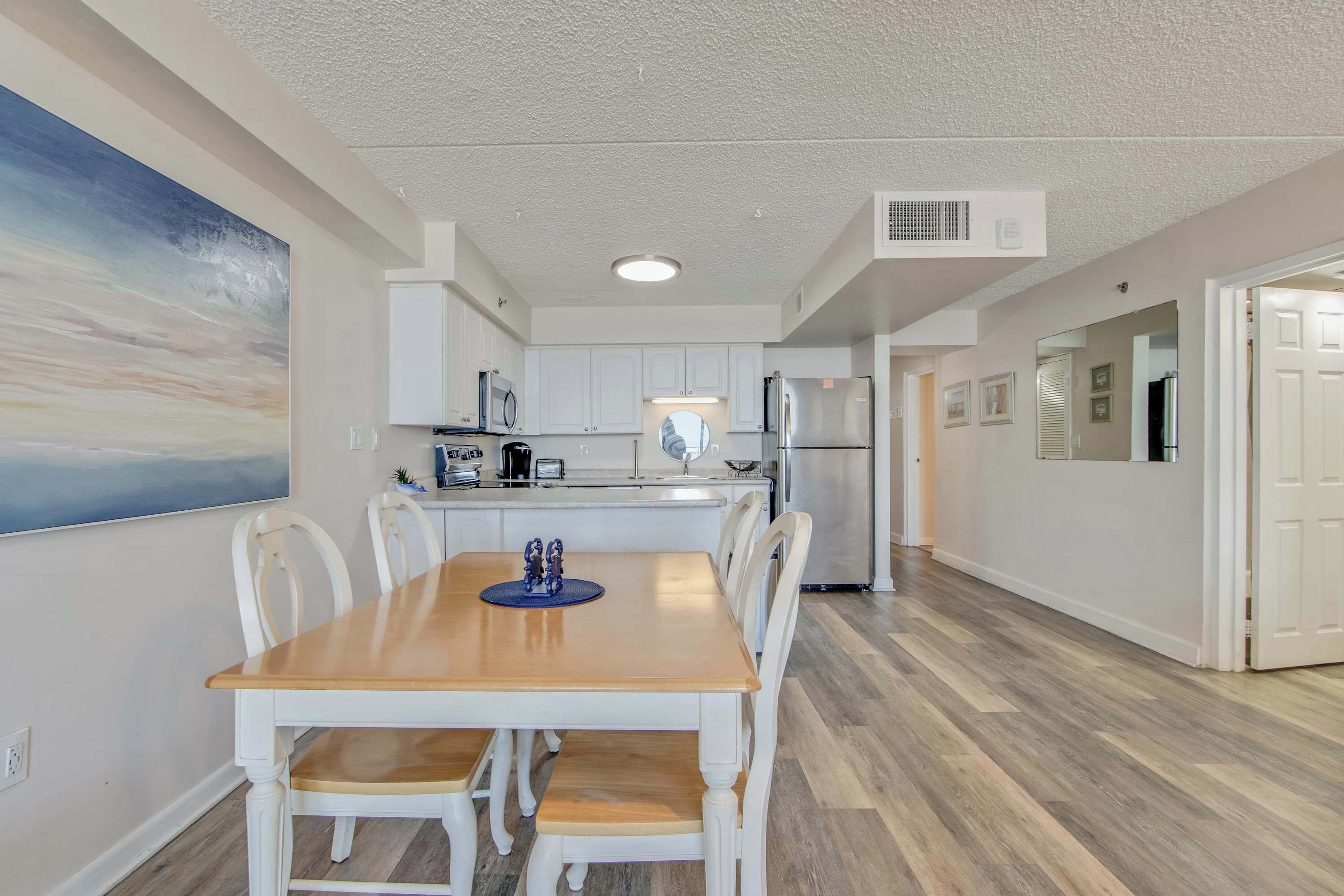 A modern kitchen with white cabinets, a wooden dining table, and a large painting on the wall.