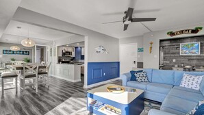 A modern living room with a blue sofa, a coffee table, and a ceiling fan. There is a kitchen area in the background with a blue accent wall. - 9400 Building #1106 (Ocean City)