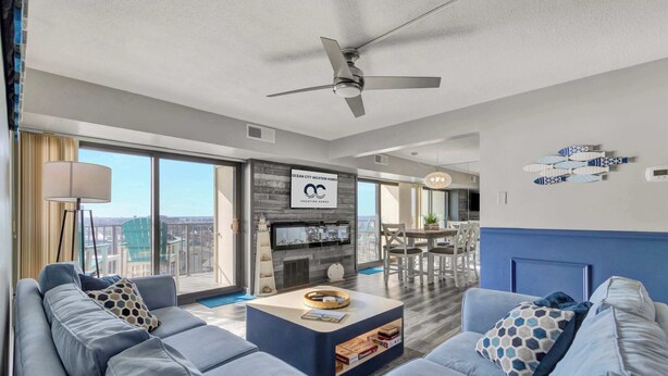 A modern living room with a large sofa, a coffee table, and a ceiling fan. There is a TV and a fireplace in the background. - 9400 Building #1106 (Ocean City)