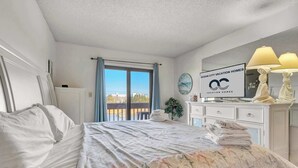 Une chambre d’hôtel avec un lit, une table de chevet, un miroir, une fenêtre avec des rideaux, une enseigne indiquant « Ocean City Vacation Homes », et une vue sur la ville.