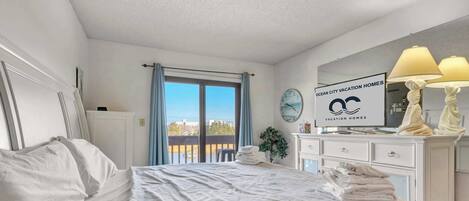 A hotel room with a bed, a nightstand, a mirror, a window with curtains, a sign for Ocean City Vacation Homes, and a view of the city.