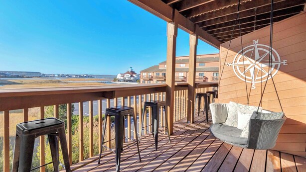 Une terrasse en bois avec un fauteuil suspendu et des tabourets de bar, offrant une vue sur la côte.
