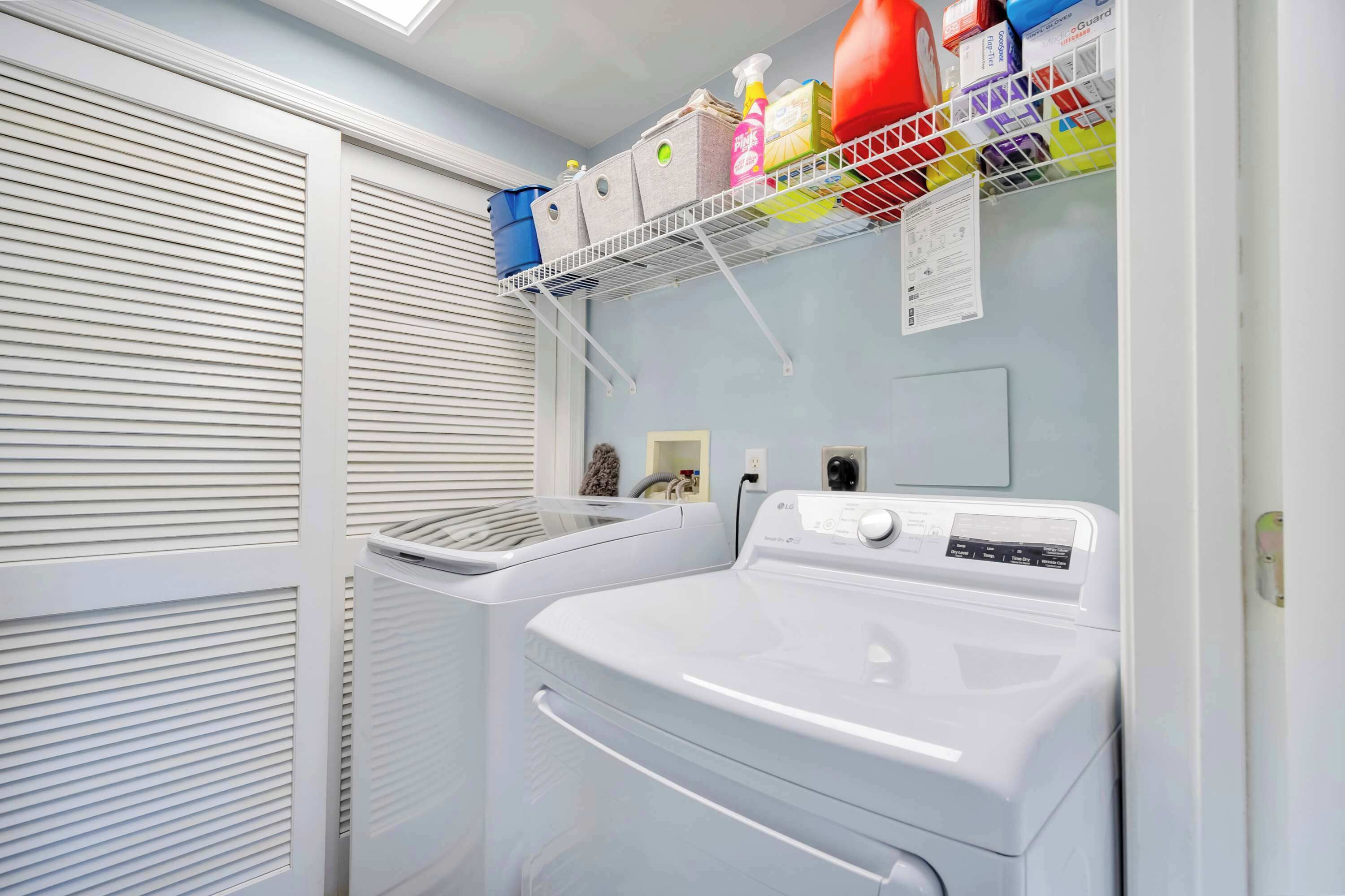 A laundry room with a front-loading washer and dryer, a shelf with cleaning supplies, and a window with blinds.