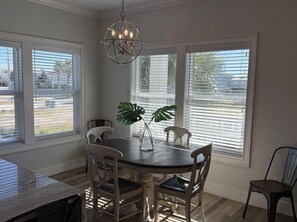 A dining area with a round table, four chairs, a chandelier, and large windows with blinds. - Lightly Salted (Gulf Shores)