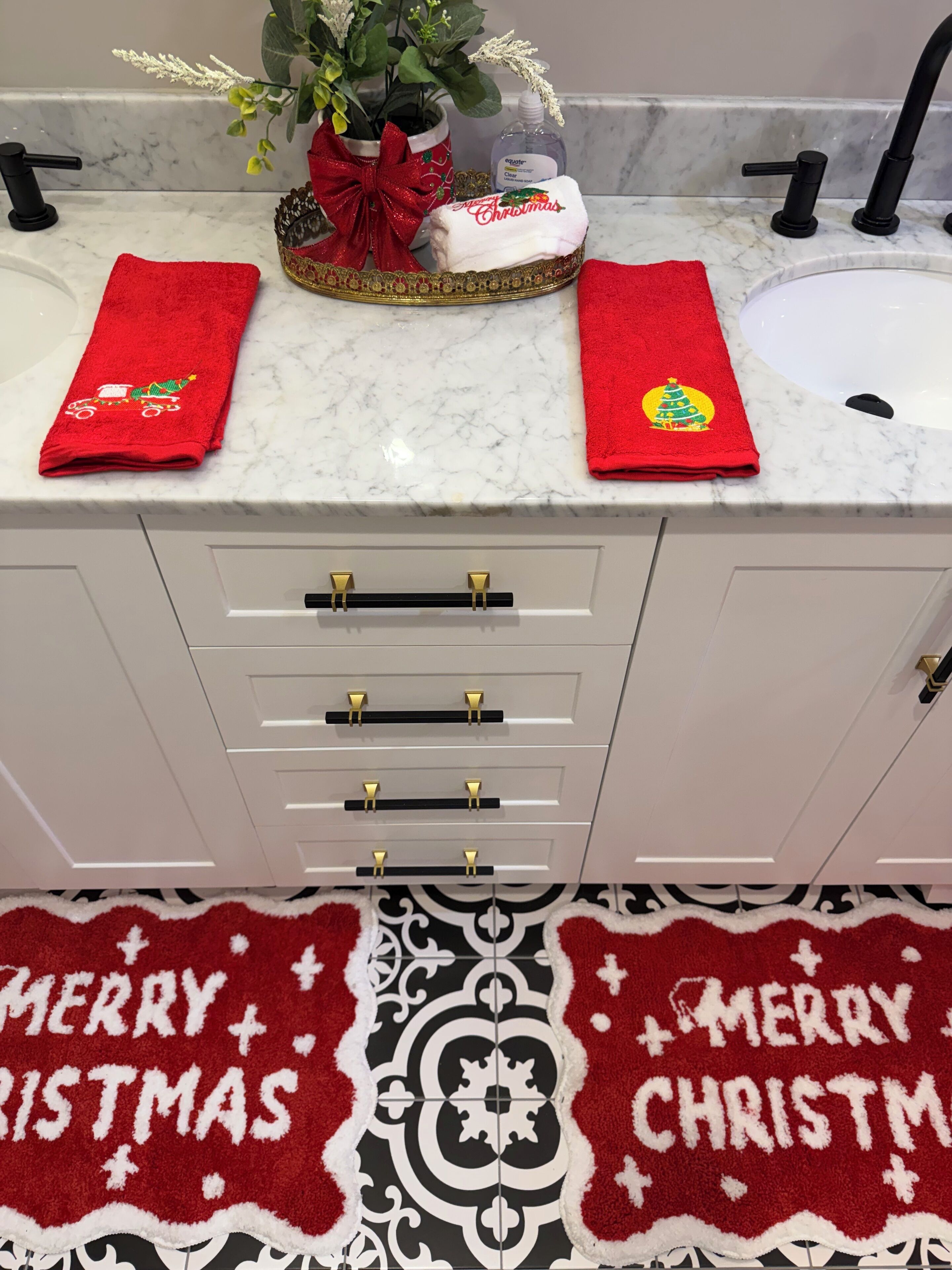 Un baño con encimera de mármol blanco, dos toallas rojas con motivos navideños, una planta decorativa y un tapete con el mensaje 'Merry Christmas'.