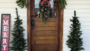 Eine Holztür mit einem Kranz, zwei Weihnachtsbäumen, einem Schild mit der Aufschrift 'MERRY' und einer Fußmatte mit der Aufschrift 'HAPPY HOUSE'.