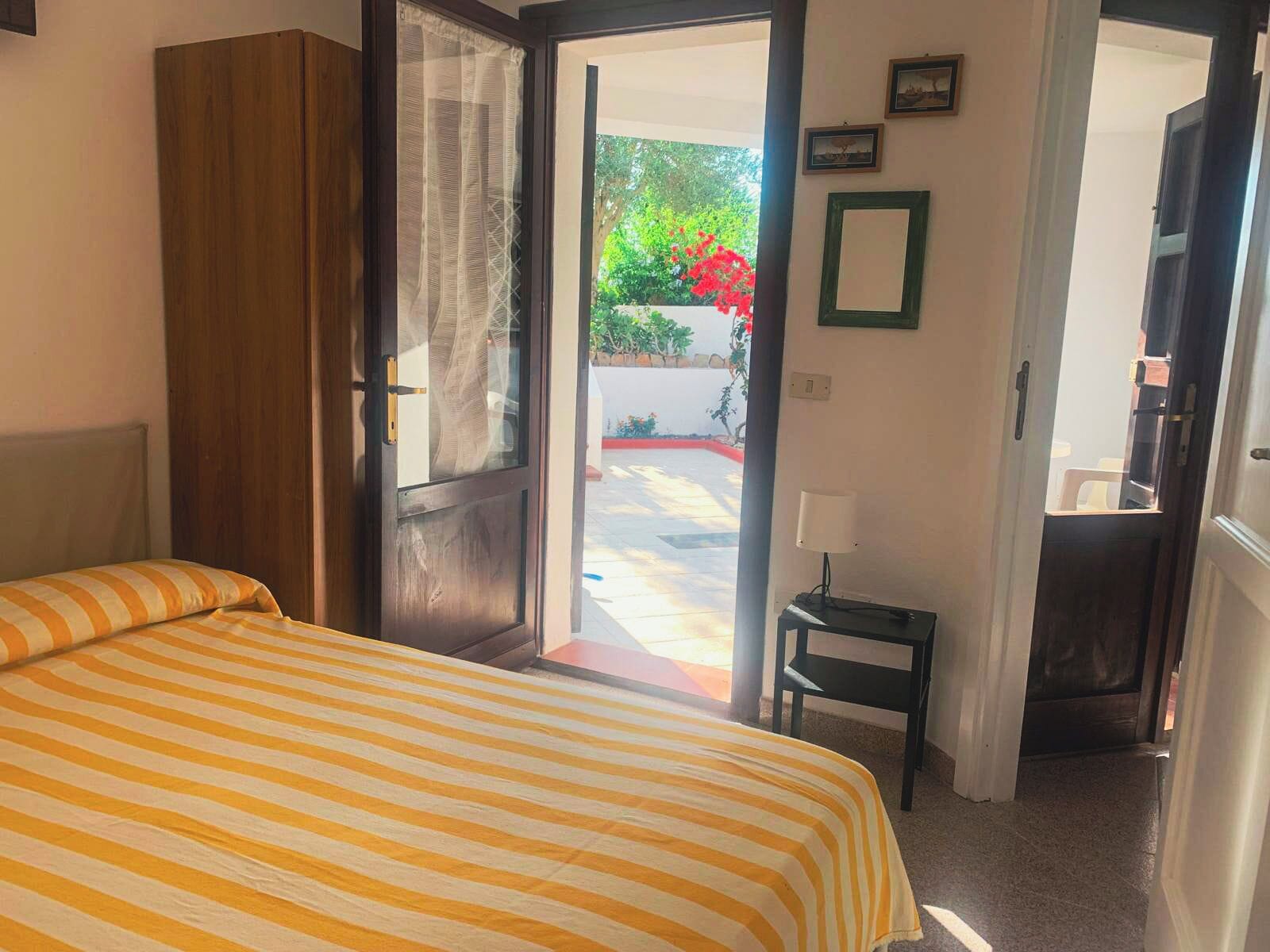 A bedroom with a bed, a wooden wardrobe, a door leading to an outdoor area with a view of greenery and a red flower, and a small table with a lamp.