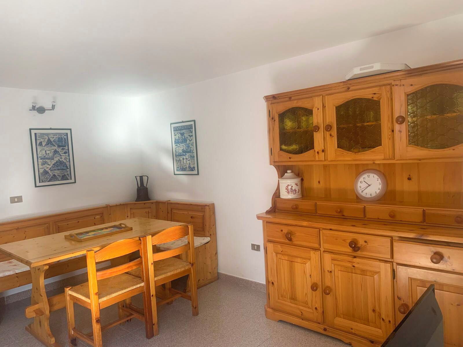 Een houten eettafel met stoelen, een houten kast met glazen deuren, en twee ingelijste foto's aan de muur.