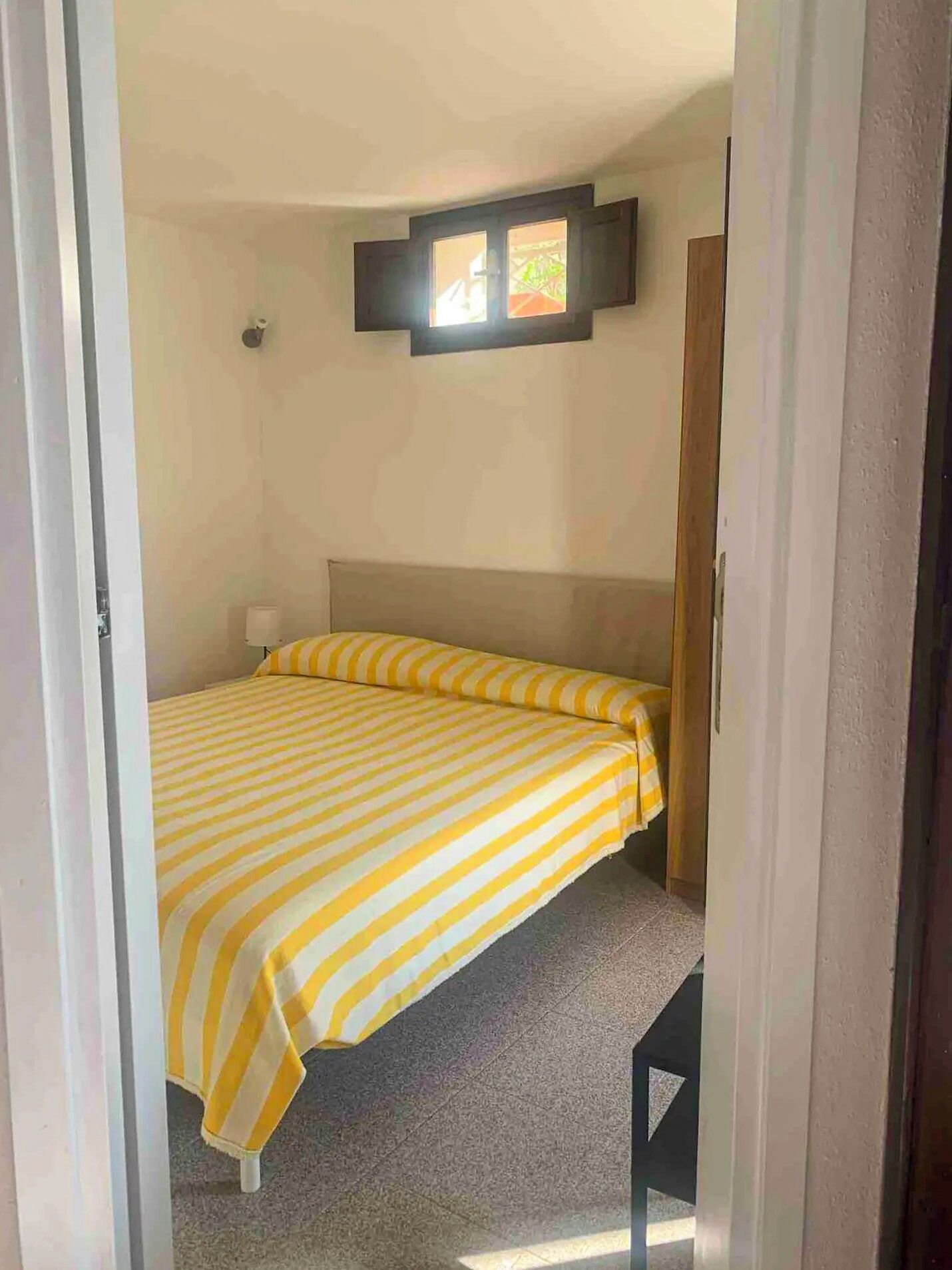 Een kleine kamer met een enkel bed, een gestreept gele en witte deken, een houten hoofdbord, een nachtkastje en een raam met uitzicht op groen.