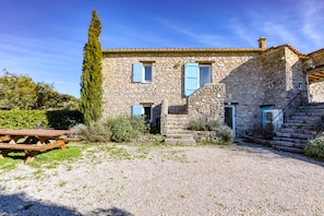 Une maison en pierre avec une allée en gravier, une table de pique-nique en bois et un grand arbre.