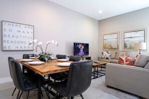 Ein modernes Wohnzimmer mit Essbereich, mit einem Holztisch, gedeckt für vier Personen, schwarzen Ledersesseln, einem grauen Sofa und zwei abstrakten Bildern an der Wand.