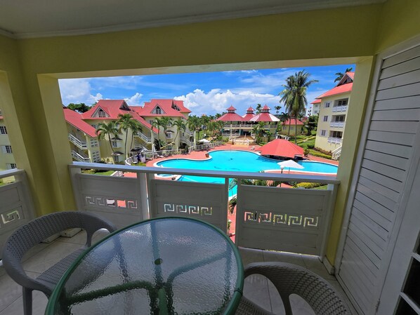 A balcony with a glass table and chairs, overlooking a swimming pool and resort buildings with red roofs. - Condo at the Mystic (Ocho Rios)
