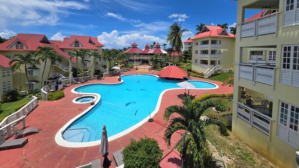 A resort with a central swimming pool, multiple buildings with red roofs, and palm trees. - Condo at the Mystic (Ocho Rios)