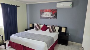 Ein Hotelzimmer mit einem Bett, Nachttischen, einer Wandklimaanlage, einem Gemälde und einem Fenster mit Vorhängen.