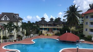 A resort with a large swimming pool, multiple buildings with red roofs, and palm trees. - Condo at the Mystic (Ocho Rios)