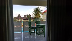 Ein Balkon mit einem Glastisch und zwei grünen Stühlen, mit Blick auf einen Pool und Gebäude mit roten Dächern.