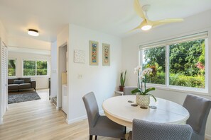 Ein modernes Wohnzimmer mit einem runden Esstisch, grauen Sesseln, einem Deckenventilator und einem großen Fenster mit Blick auf Grünflächen.