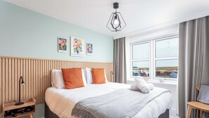 A neatly arranged bedroom with a large bed, a bedside table, a lamp, and a view of the outdoors through a window. - Old Prestwick Point (Prestwick)