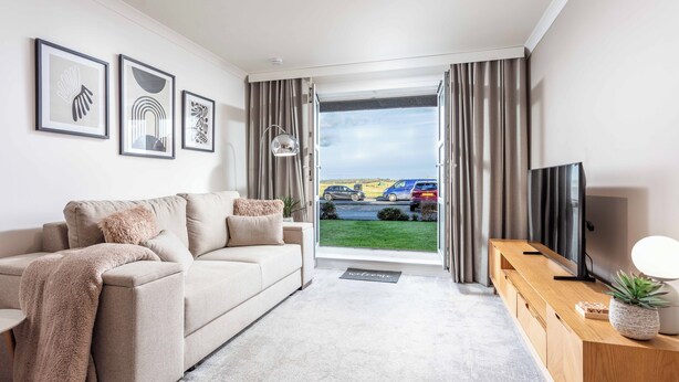 A modern living room with a sofa, a coffee table, a TV on a wooden stand, and a view of a parking lot through large windows. - Old Prestwick Point (Prestwick)