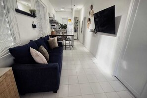 A modern living room with a dark blue sofa, a dining area with a table and chairs, and a TV mounted on the wall. - Casa Qodesh #1: Oasis Eco-Gem, Solar & Water (Arecibo)