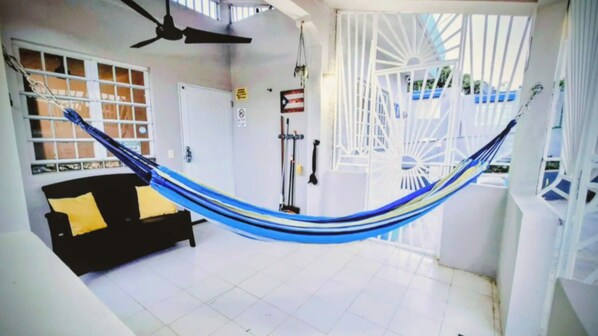 A hammock in a room with a black sofa, a ceiling fan, and a white door. - Casa Qodesh #1: Oasis Eco-Gem, Solar & Water (Arecibo)